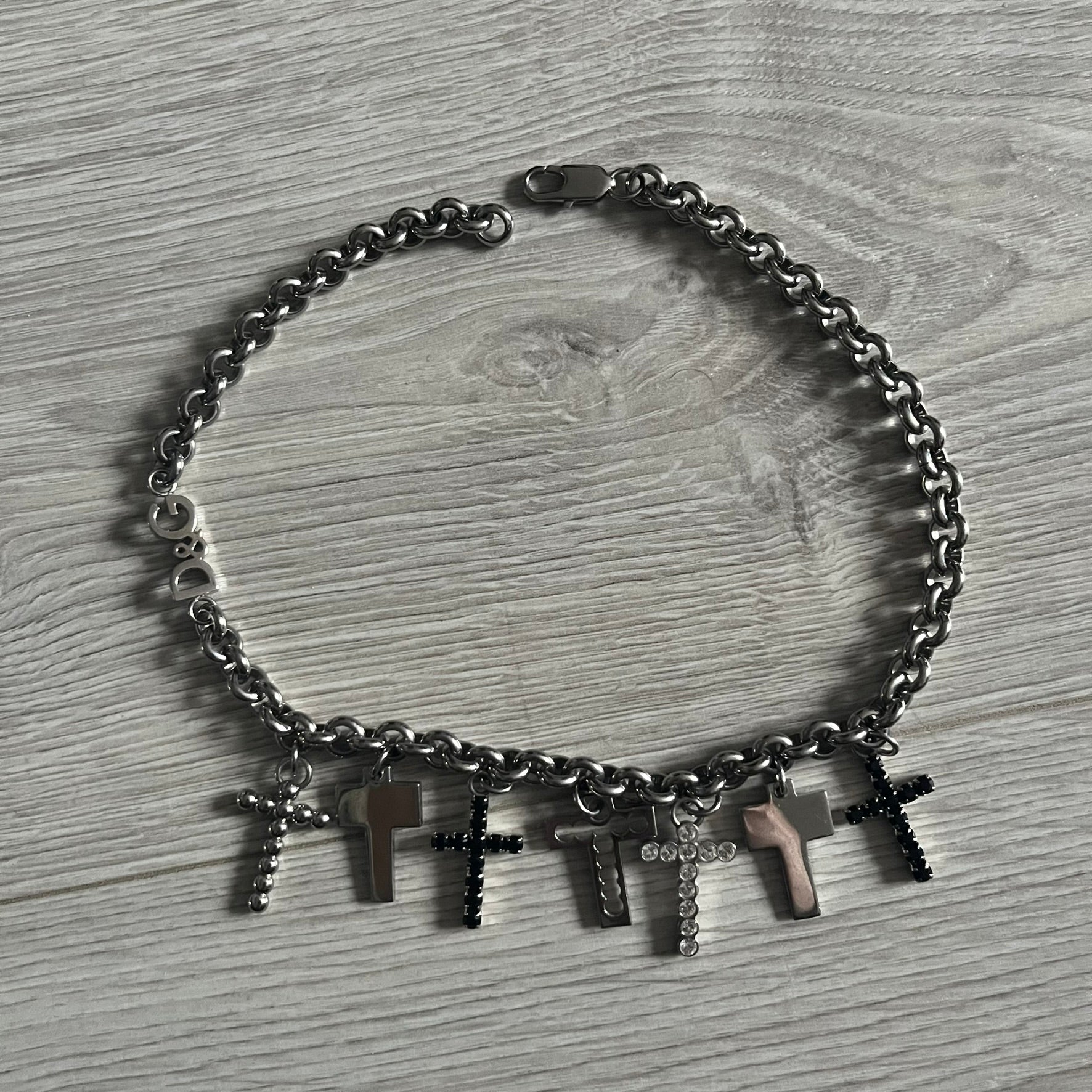 Dolce & Gabbana 7 Crosses Stainless Steel Choker Necklace. Crafted from polished stainless steel, this necklace features seven dangling crosses in varying designs, including full metal, black stones, white stones, and cut-out styles. One section of the chain incorporates a D&G logo in place of links. Comes with original box and dustbag.