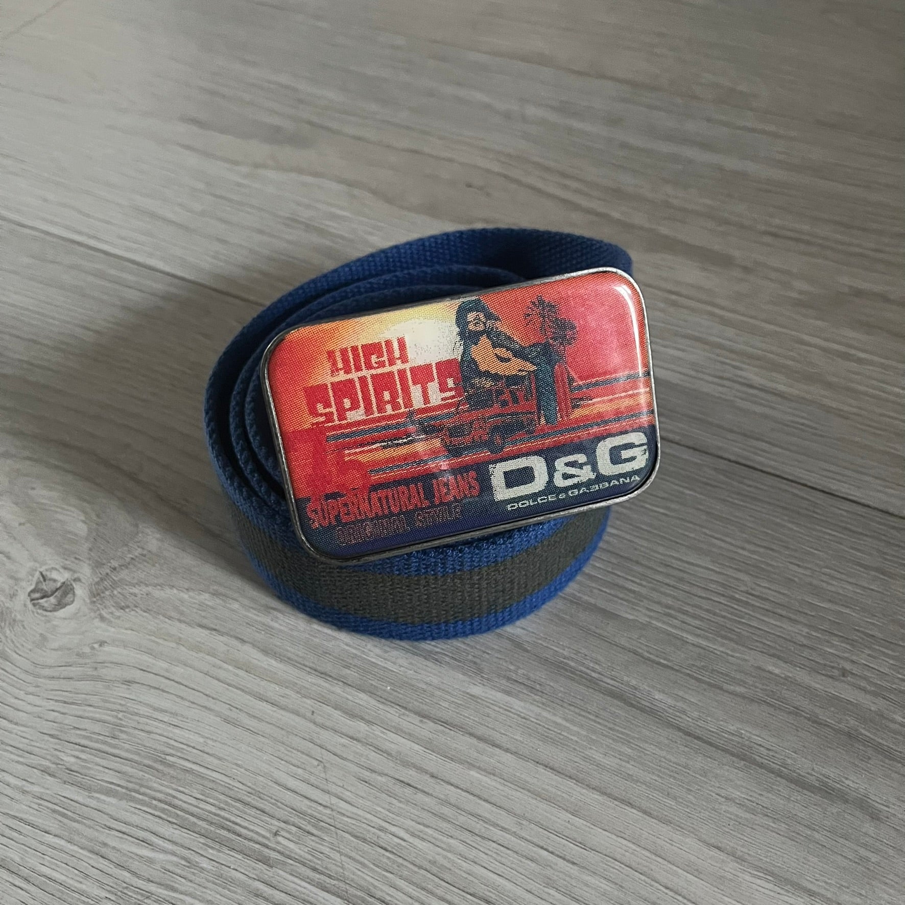 Dolce &amp; Gabbana “High Spirits” Fabric Belt in blue with a brown/gray central stripe and enamel-finished metal buckle. The buckle features vintage iconography and “High Spirits” lettering in vivid orange/red and navy tones. A rare 2000s-era Dolce &amp; Gabbana belt showcasing the brand’s early denim aesthetic.