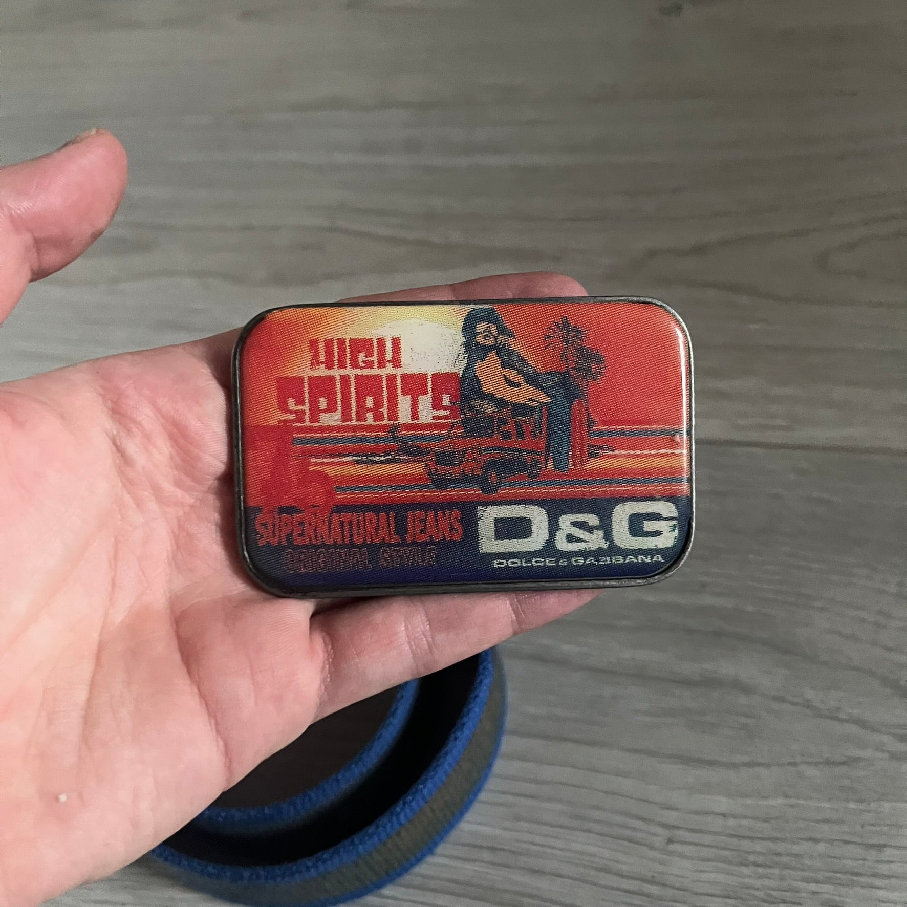 Dolce &amp; Gabbana “High Spirits” Fabric Belt in blue with a brown/gray central stripe and enamel-finished metal buckle. The buckle features vintage iconography and “High Spirits” lettering in vivid orange/red and navy tones. A rare 2000s-era Dolce &amp; Gabbana belt showcasing the brand’s early denim aesthetic.