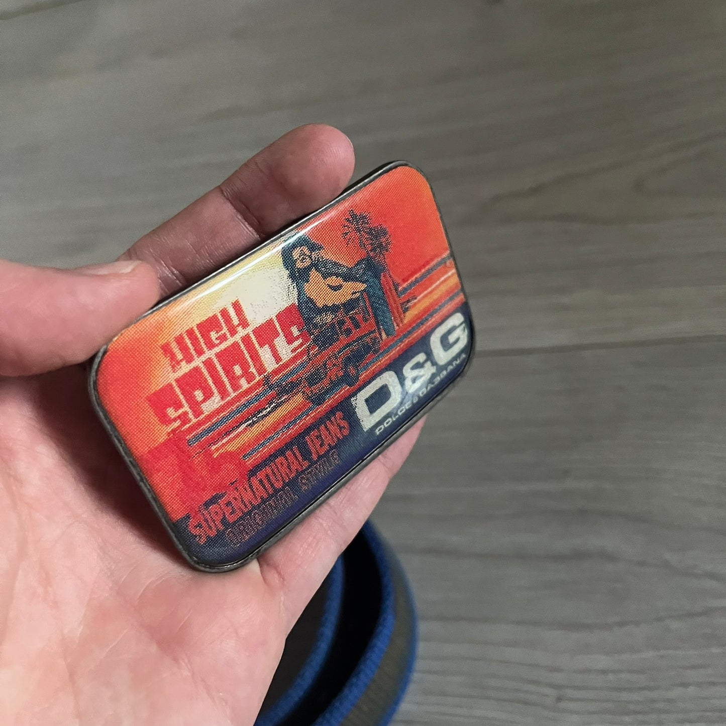 Dolce &amp; Gabbana “High Spirits” Fabric Belt in blue with a brown/gray central stripe and enamel-finished metal buckle. The buckle features vintage iconography and “High Spirits” lettering in vivid orange/red and navy tones. A rare 2000s-era Dolce &amp; Gabbana belt showcasing the brand’s early denim aesthetic.