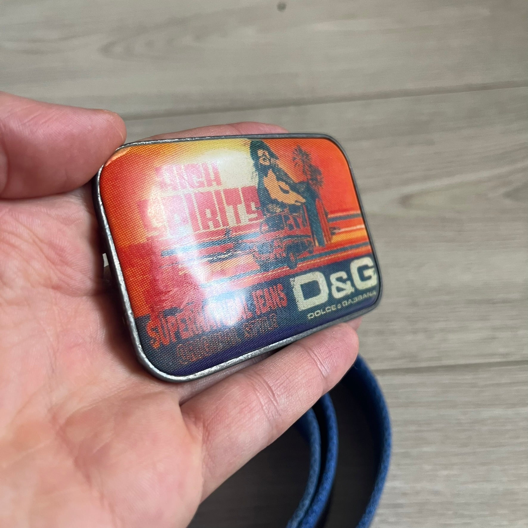 Dolce &amp; Gabbana “High Spirits” Fabric Belt in blue with a brown central stripe and enamel-finished metal buckle. The buckle features vintage iconography and “High Spirits” lettering in vivid orange/red and navy tones. A rare 2000s-era Dolce &amp; Gabbana belt showcasing the brand’s early denim aesthetic.