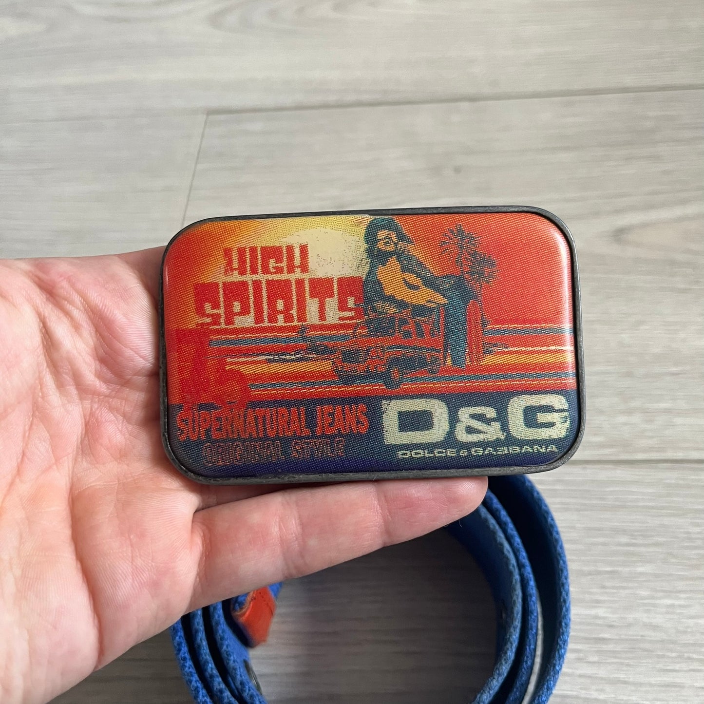 Dolce &amp; Gabbana “High Spirits” Fabric Belt in blue with a brown central stripe and enamel-finished metal buckle. The buckle features vintage iconography and “High Spirits” lettering in vivid orange/red and navy tones. A rare 2000s-era Dolce &amp; Gabbana belt showcasing the brand’s early denim aesthetic.