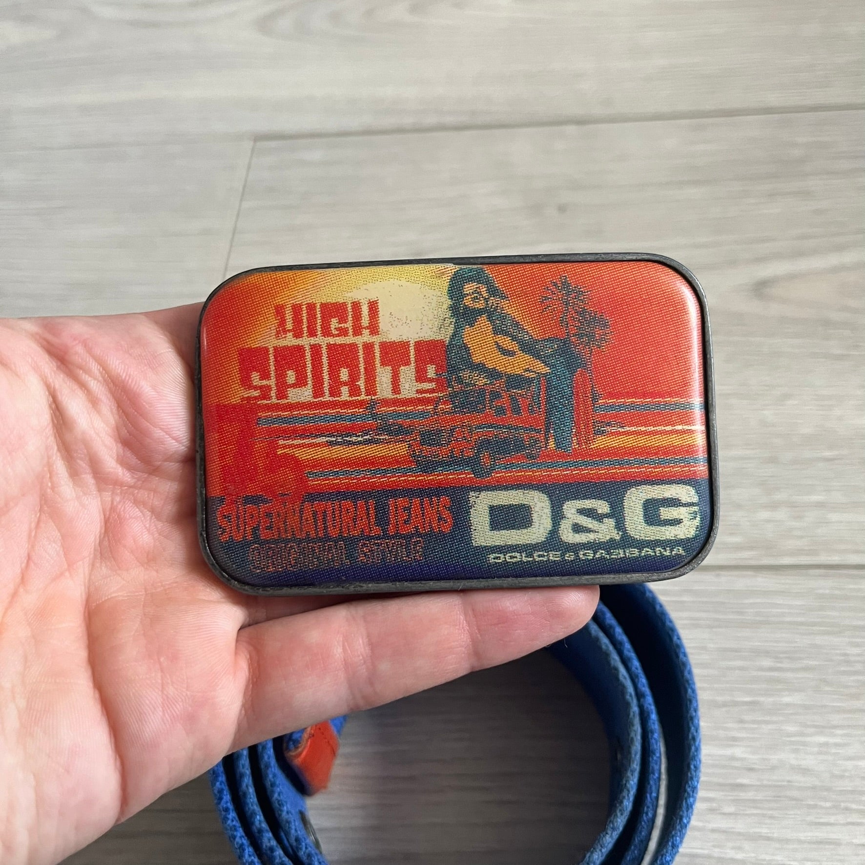 Dolce &amp; Gabbana “High Spirits” Fabric Belt in blue with a brown central stripe and enamel-finished metal buckle. The buckle features vintage iconography and “High Spirits” lettering in vivid orange/red and navy tones. A rare 2000s-era Dolce &amp; Gabbana belt showcasing the brand’s early denim aesthetic.