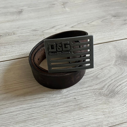 Dolce &amp; Gabbana USA Flag Leather Belt crafted from brown leather with concave, sunken Dolce &amp; Gabbana and D&amp;G text detailing along the strap. The metal buckle is designed in a flag-inspired layout, featuring six horizontal cut-out lines and D&amp;G Dolce &amp; Gabbana text in the upper-left corner in place of traditional stars. The belt shows visible wear consistent with age, giving it a heavily worn, vintage finish.