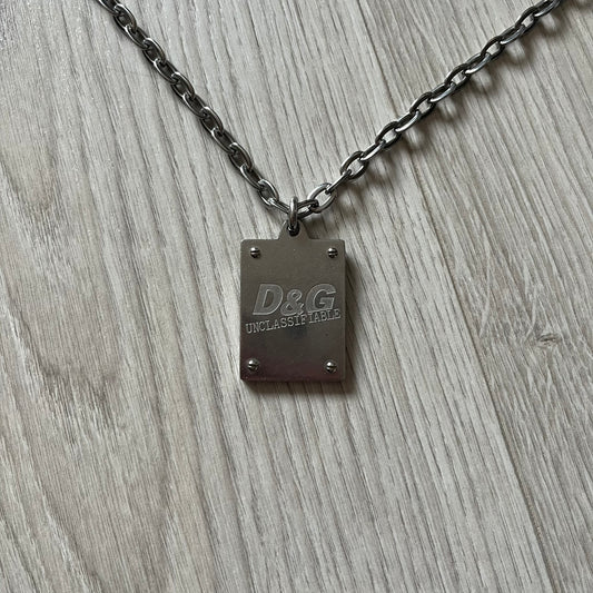 Dolce & Gabbana 'Unclassifiable' Dog Tag Necklace in stainless steel with a silver cable chain and lobster clasp. Features a thick dog tag with four visible screws on the front and engraved D& G logo with “Unclassifiable” diagonally beneath.