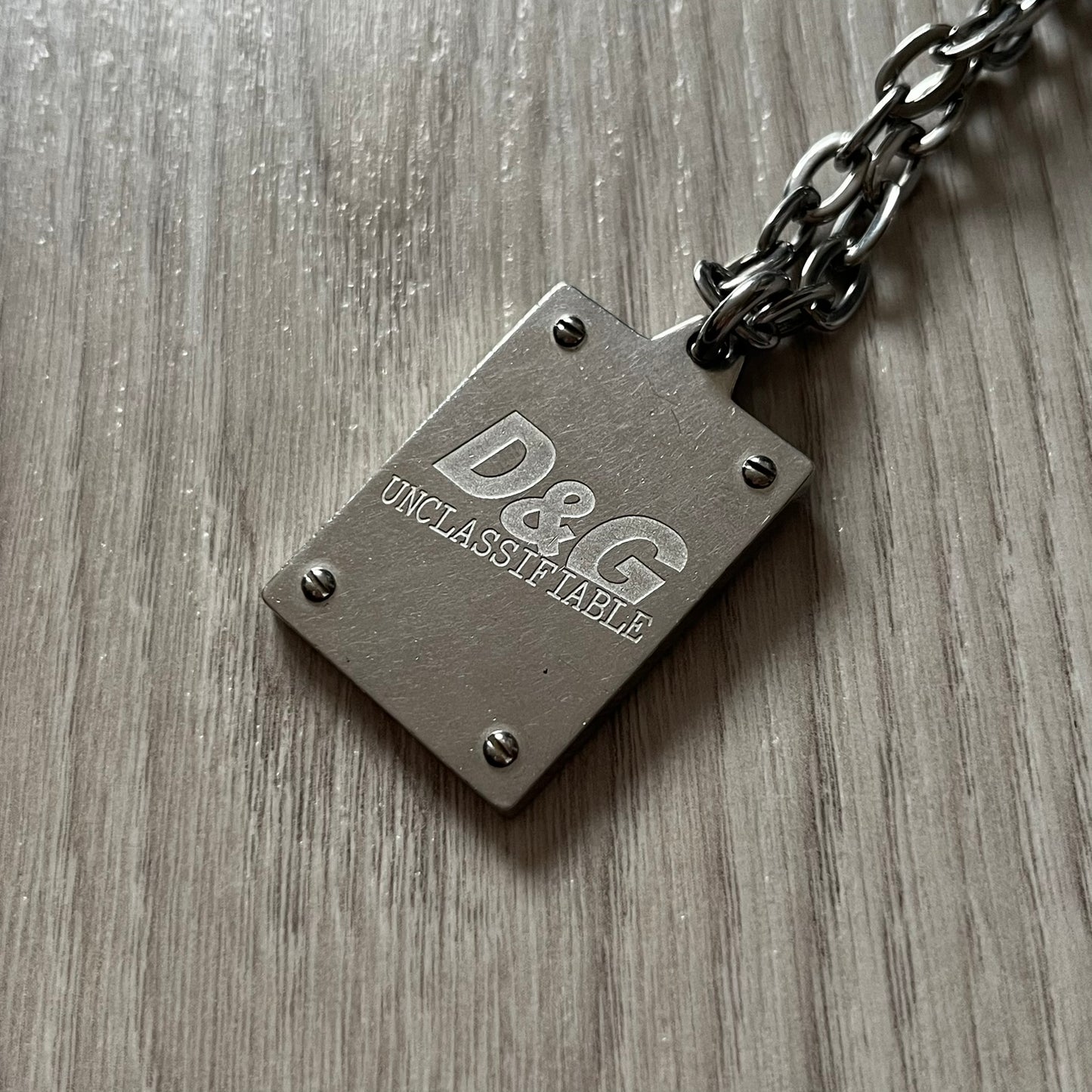 Dolce & Gabbana 'Unclassifiable' Dog Tag Necklace in stainless steel with a silver cable chain and lobster clasp. Features a thick dog tag with four visible screws on the front and engraved D& G logo with “Unclassifiable” diagonally beneath.