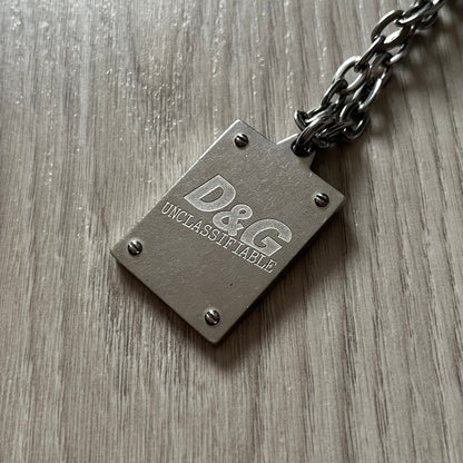 Dolce & Gabbana 'Unclassifiable' Dog Tag Necklace in stainless steel with a silver cable chain and lobster clasp. Features a thick dog tag with four visible screws on the front and engraved D& G logo with “Unclassifiable” diagonally beneath.