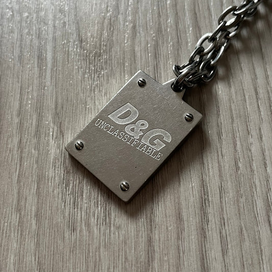 Dolce & Gabbana 'Unclassifiable' Dog Tag Necklace in stainless steel with a silver cable chain and lobster clasp. Features a thick dog tag with four visible screws on the front and engraved D& G logo with “Unclassifiable” diagonally beneath.
