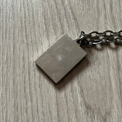 Dolce & Gabbana 'Unclassifiable' Dog Tag Necklace in stainless steel with a silver cable chain and lobster clasp. Features a thick dog tag with four visible screws on the front and engraved D& G logo with “Unclassifiable” diagonally beneath.