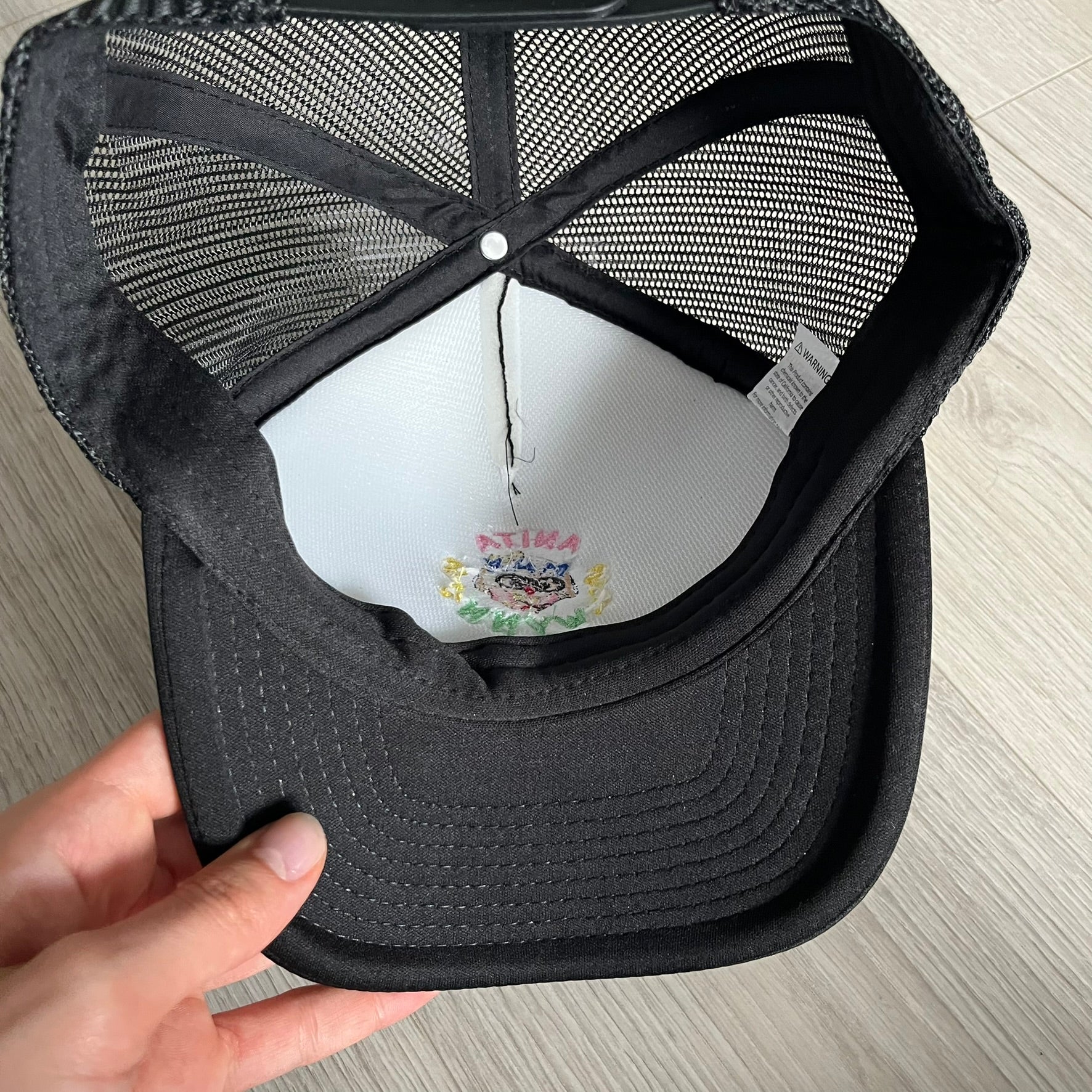 Drake Anita Max Wynn Pink Trucker Hat in all-black front panel, sides and cap, featuring a collection of graphics including a woman with an oversized head, two yellow dollar signs, and “ANITA” in pink, “MAX” in blue, and “WYNN” in green. Designed with a mesh back for breathability, a snapback closure for adjustability, and polyester construction to keep the head cool. A playful tribute to Drake’s alter ego and love of g4mbling.