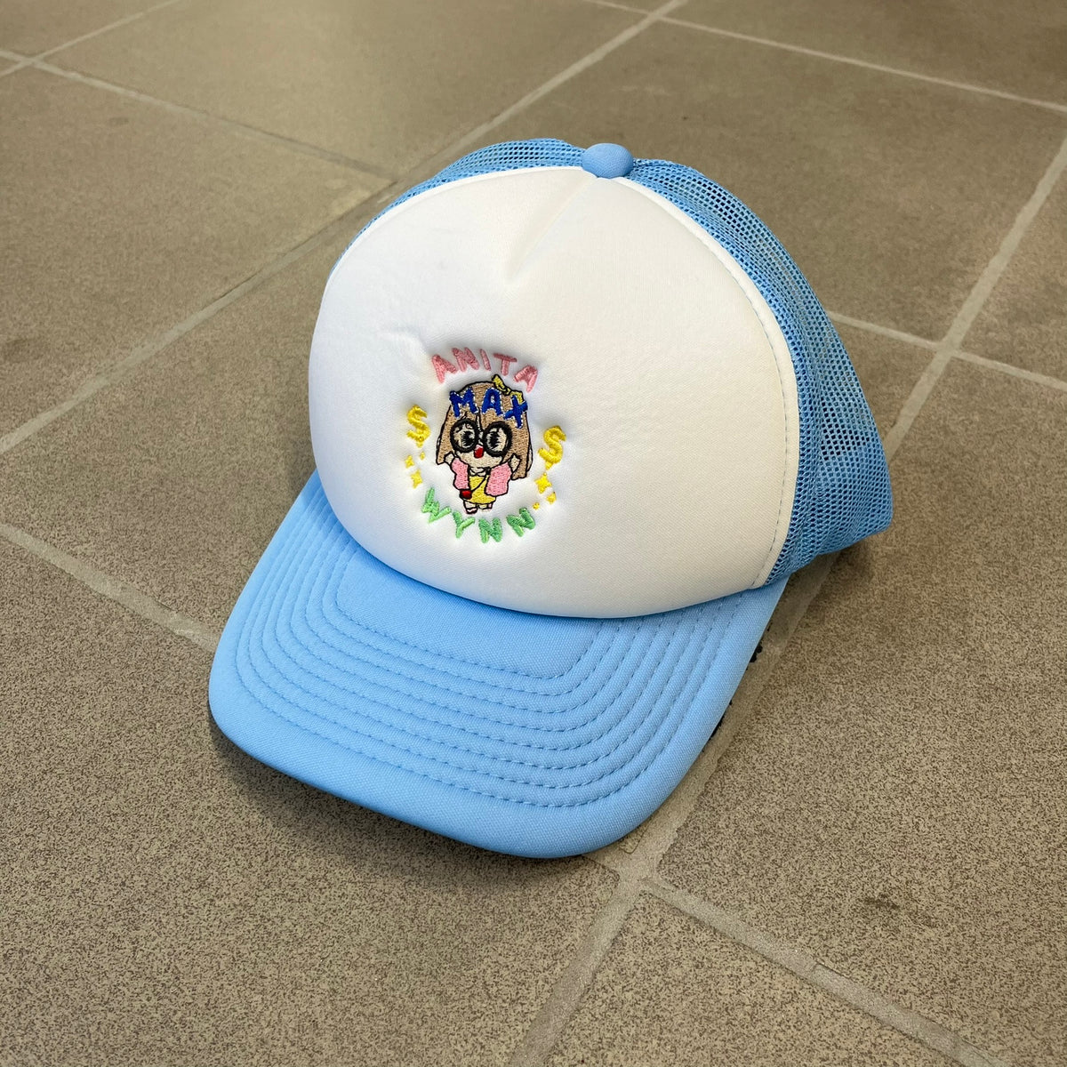 Drake Anita Max Wynn Blue Trucker Cap with a white front panel and baby blue sides and cap, featuring a collection of graphics including a woman with an oversized head, two yellow dollar signs, and “ANITA” in pink, “MAX” in blue, and “WYNN” in green. Designed with a mesh back for breathability, a snapback closure for adjustability, and polyester construction to keep the head cool.