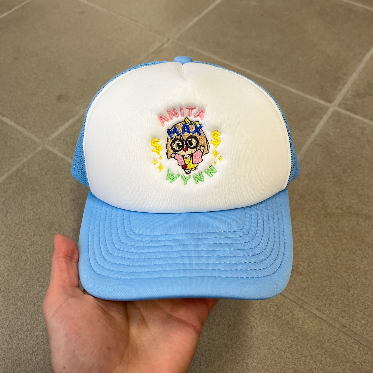 Drake Anita Max Wynn Blue Trucker Cap with a white front panel and baby blue sides and cap, featuring a collection of graphics including a woman with an oversized head, two yellow dollar signs, and “ANITA” in pink, “MAX” in blue, and “WYNN” in green. Designed with a mesh back for breathability, a snapback closure for adjustability, and polyester construction to keep the head cool.