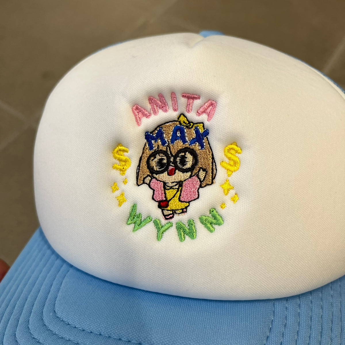 Drake Anita Max Wynn Blue Trucker Cap with a white front panel and baby blue sides and cap, featuring a collection of graphics including a woman with an oversized head, two yellow dollar signs, and “ANITA” in pink, “MAX” in blue, and “WYNN” in green. Designed with a mesh back for breathability, a snapback closure for adjustability, and polyester construction to keep the head cool.