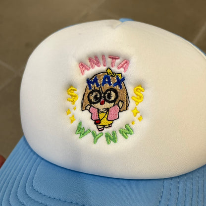 Drake Anita Max Wynn Blue Trucker Cap with a white front panel and baby blue sides and cap, featuring a collection of graphics including a woman with an oversized head, two yellow dollar signs, and “ANITA” in pink, “MAX” in blue, and “WYNN” in green. Designed with a mesh back for breathability, a snapback closure for adjustability, and polyester construction to keep the head cool.