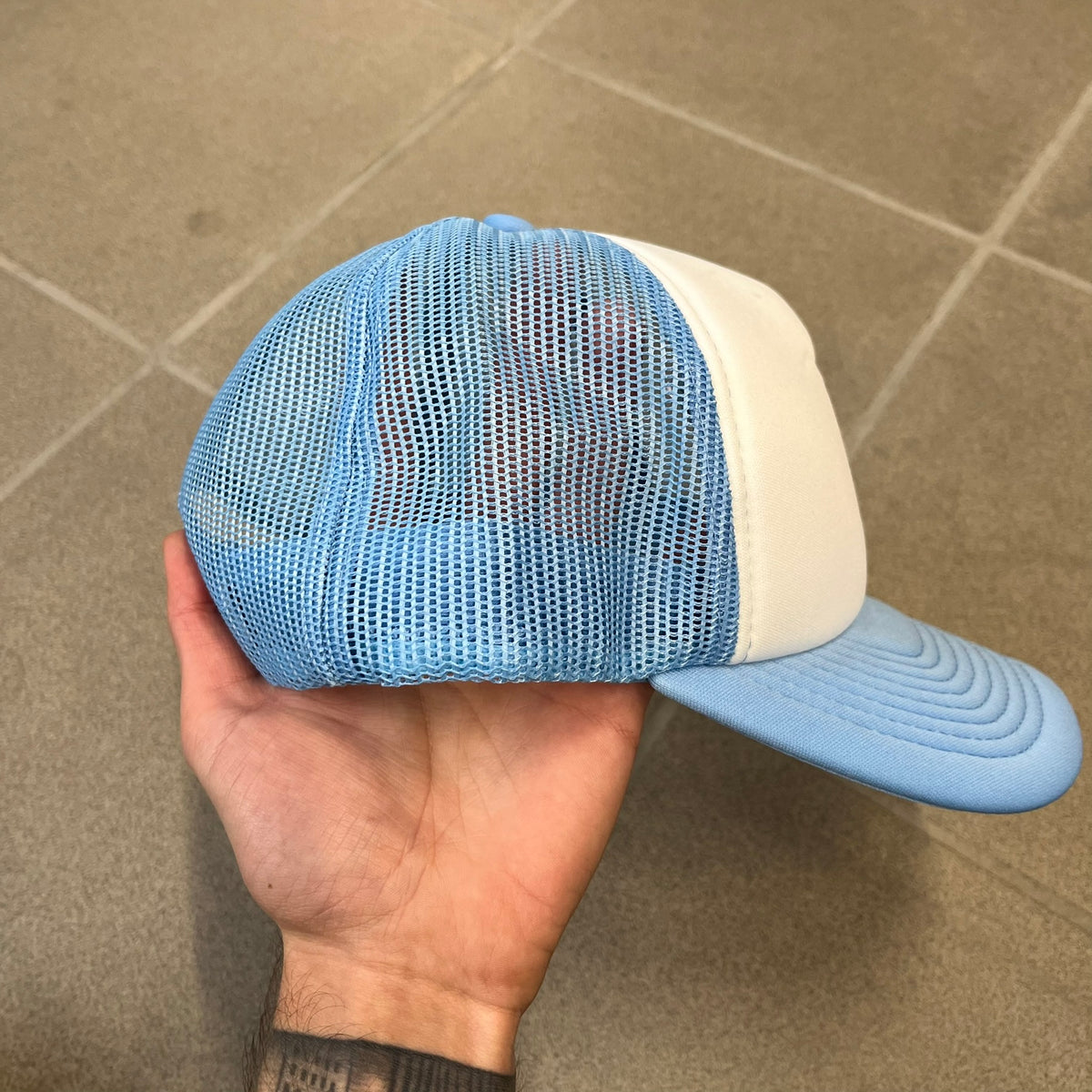 Drake Anita Max Wynn Blue Trucker Cap with a white front panel and baby blue sides and cap, featuring a collection of graphics including a woman with an oversized head, two yellow dollar signs, and “ANITA” in pink, “MAX” in blue, and “WYNN” in green. Designed with a mesh back for breathability, a snapback closure for adjustability, and polyester construction to keep the head cool.