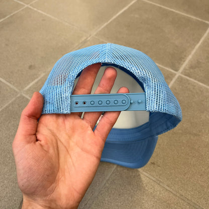 Drake Anita Max Wynn Blue Trucker Cap with a white front panel and baby blue sides and cap, featuring a collection of graphics including a woman with an oversized head, two yellow dollar signs, and “ANITA” in pink, “MAX” in blue, and “WYNN” in green. Designed with a mesh back for breathability, a snapback closure for adjustability, and polyester construction to keep the head cool.