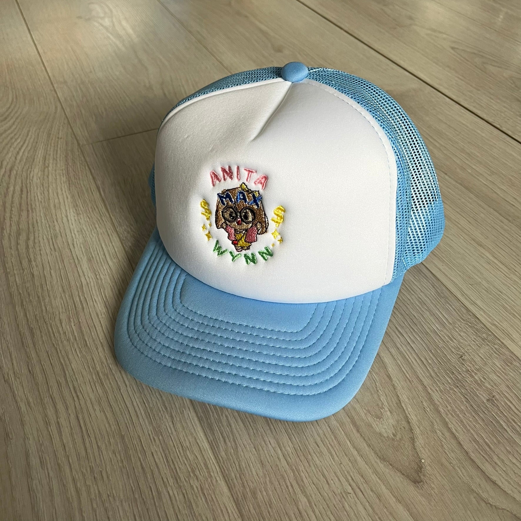 Drake Anita Max Wynn Pink Trucker Hat with a white front panel and baby blue sides and cap, featuring a collection of graphics including a woman with an oversized head, two yellow dollar signs, and “ANITA” in pink, “MAX” in blue, and “WYNN” in green. Designed with a mesh back for breathability, a snapback closure for adjustability, and polyester construction to keep the head cool. A playful tribute to Drake’s alter ego and love of g4mbling.