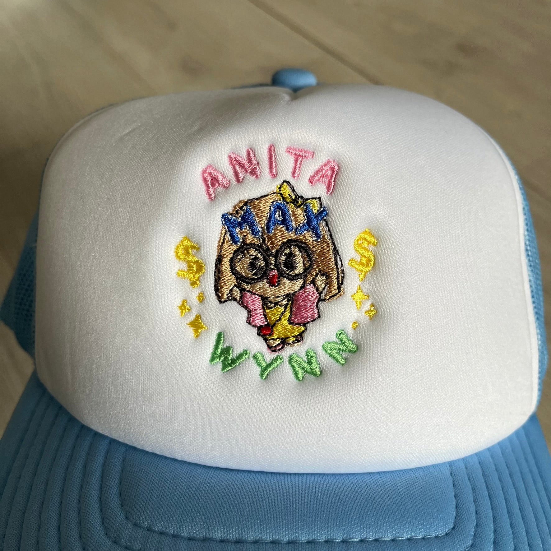 Drake Anita Max Wynn Pink Trucker Hat with a white front panel and baby blue sides and cap, featuring a collection of graphics including a woman with an oversized head, two yellow dollar signs, and “ANITA” in pink, “MAX” in blue, and “WYNN” in green. Designed with a mesh back for breathability, a snapback closure for adjustability, and polyester construction to keep the head cool. A playful tribute to Drake’s alter ego and love of g4mbling.