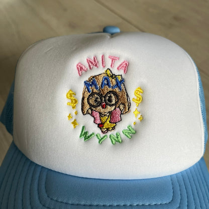 Drake Anita Max Wynn Pink Trucker Hat with a white front panel and baby blue sides and cap, featuring a collection of graphics including a woman with an oversized head, two yellow dollar signs, and “ANITA” in pink, “MAX” in blue, and “WYNN” in green. Designed with a mesh back for breathability, a snapback closure for adjustability, and polyester construction to keep the head cool. A playful tribute to Drake’s alter ego and love of g4mbling.
