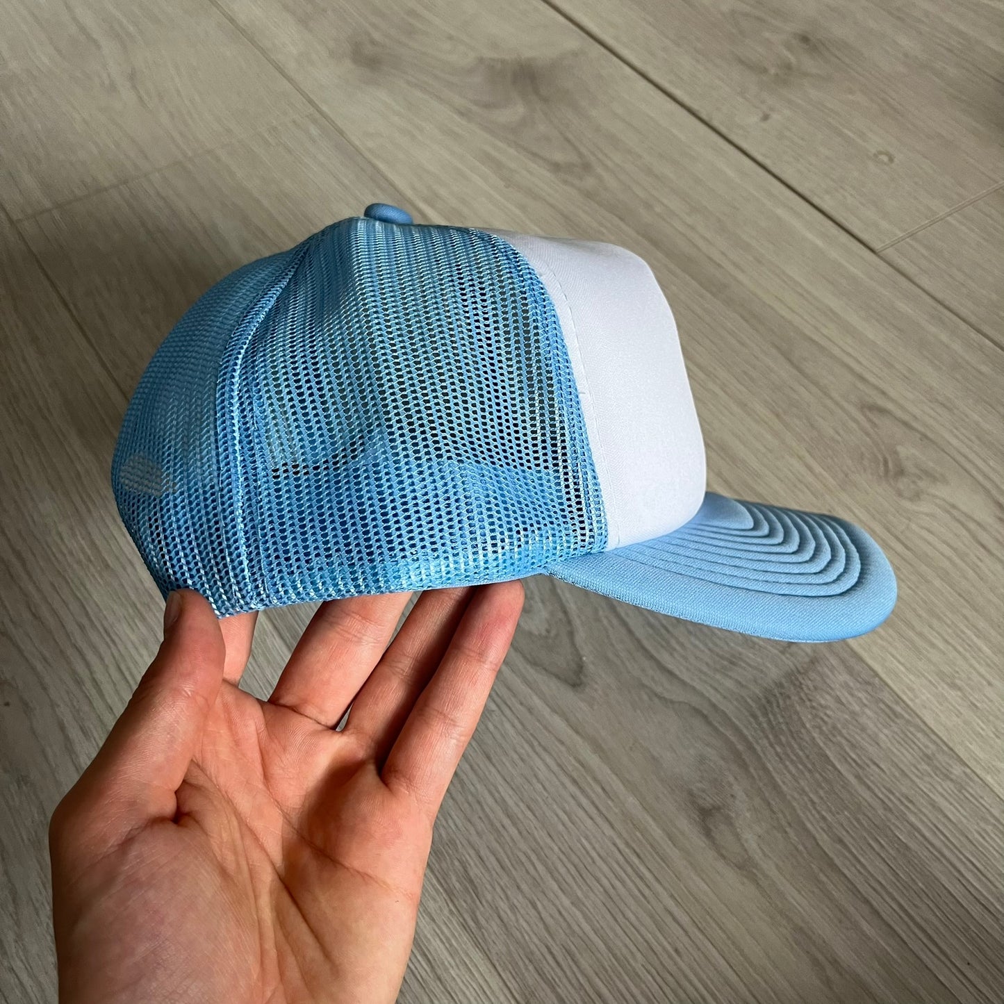 Drake Anita Max Wynn Pink Trucker Hat with a white front panel and baby blue sides and cap, featuring a collection of graphics including a woman with an oversized head, two yellow dollar signs, and “ANITA” in pink, “MAX” in blue, and “WYNN” in green. Designed with a mesh back for breathability, a snapback closure for adjustability, and polyester construction to keep the head cool. A playful tribute to Drake’s alter ego and love of g4mbling.