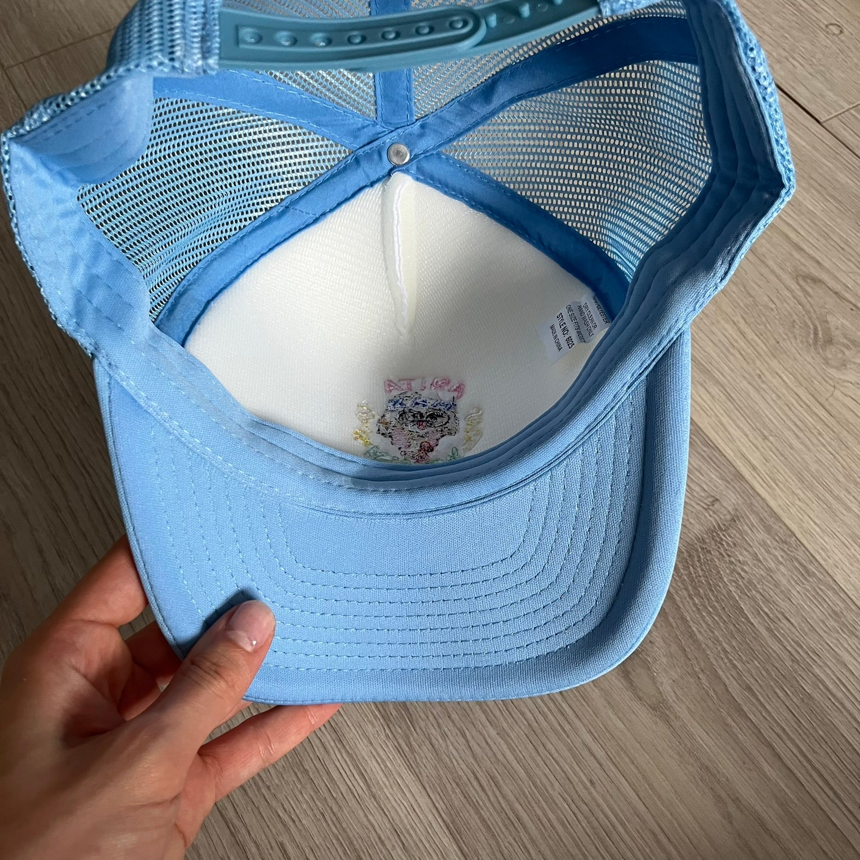 Drake Anita Max Wynn Pink Trucker Hat with a white front panel and baby blue sides and cap, featuring a collection of graphics including a woman with an oversized head, two yellow dollar signs, and “ANITA” in pink, “MAX” in blue, and “WYNN” in green. Designed with a mesh back for breathability, a snapback closure for adjustability, and polyester construction to keep the head cool. A playful tribute to Drake’s alter ego and love of g4mbling.
