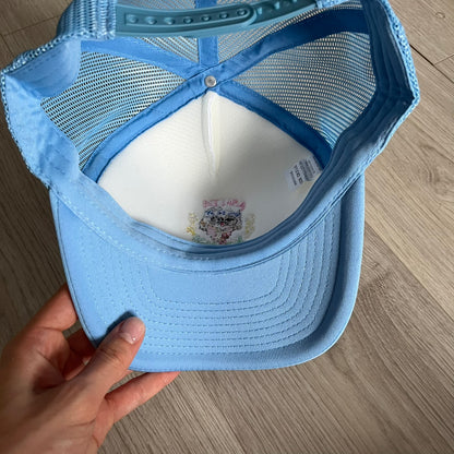 Drake Anita Max Wynn Pink Trucker Hat with a white front panel and baby blue sides and cap, featuring a collection of graphics including a woman with an oversized head, two yellow dollar signs, and “ANITA” in pink, “MAX” in blue, and “WYNN” in green. Designed with a mesh back for breathability, a snapback closure for adjustability, and polyester construction to keep the head cool. A playful tribute to Drake’s alter ego and love of g4mbling.