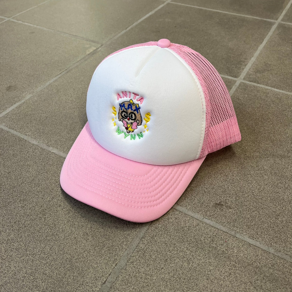 Drake Anita Max Wynn Pink Trucker Cap with a white front panel and baby blue sides and cap, featuring a collection of graphics including a woman with an oversized head, two yellow dollar signs, and “ANITA” in pink, “MAX” in blue, and “WYNN” in green. Designed with a mesh back for breathability, a snapback closure for adjustability, and polyester construction to keep the head cool.