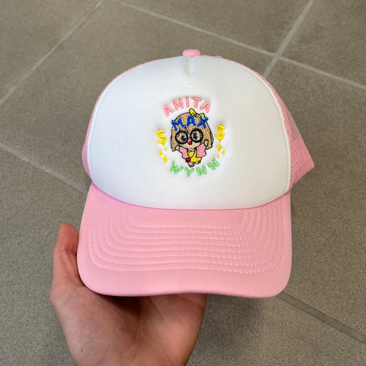 Drake Anita Max Wynn Pink Trucker Cap with a white front panel and baby blue sides and cap, featuring a collection of graphics including a woman with an oversized head, two yellow dollar signs, and “ANITA” in pink, “MAX” in blue, and “WYNN” in green. Designed with a mesh back for breathability, a snapback closure for adjustability, and polyester construction to keep the head cool.