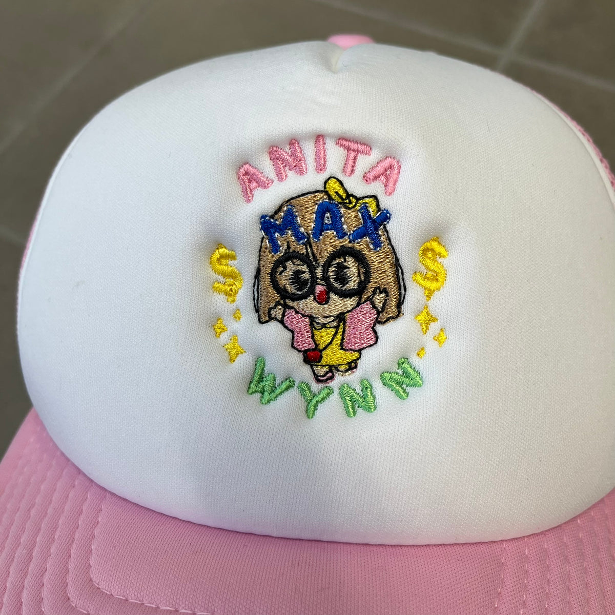 Drake Anita Max Wynn Pink Trucker Cap with a white front panel and baby blue sides and cap, featuring a collection of graphics including a woman with an oversized head, two yellow dollar signs, and “ANITA” in pink, “MAX” in blue, and “WYNN” in green. Designed with a mesh back for breathability, a snapback closure for adjustability, and polyester construction to keep the head cool.