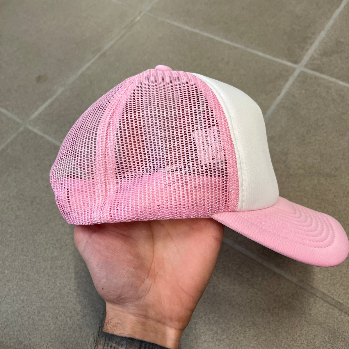 Drake Anita Max Wynn Pink Trucker Cap with a white front panel and baby blue sides and cap, featuring a collection of graphics including a woman with an oversized head, two yellow dollar signs, and “ANITA” in pink, “MAX” in blue, and “WYNN” in green. Designed with a mesh back for breathability, a snapback closure for adjustability, and polyester construction to keep the head cool.