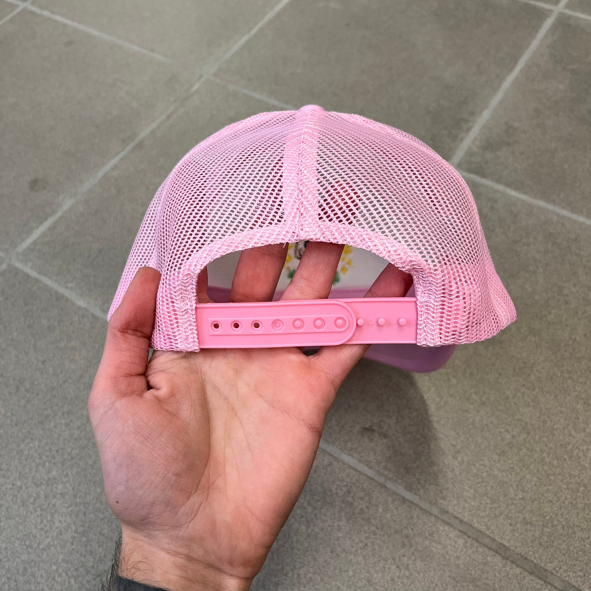 Drake Anita Max Wynn Pink Trucker Cap with a white front panel and baby blue sides and cap, featuring a collection of graphics including a woman with an oversized head, two yellow dollar signs, and “ANITA” in pink, “MAX” in blue, and “WYNN” in green. Designed with a mesh back for breathability, a snapback closure for adjustability, and polyester construction to keep the head cool.