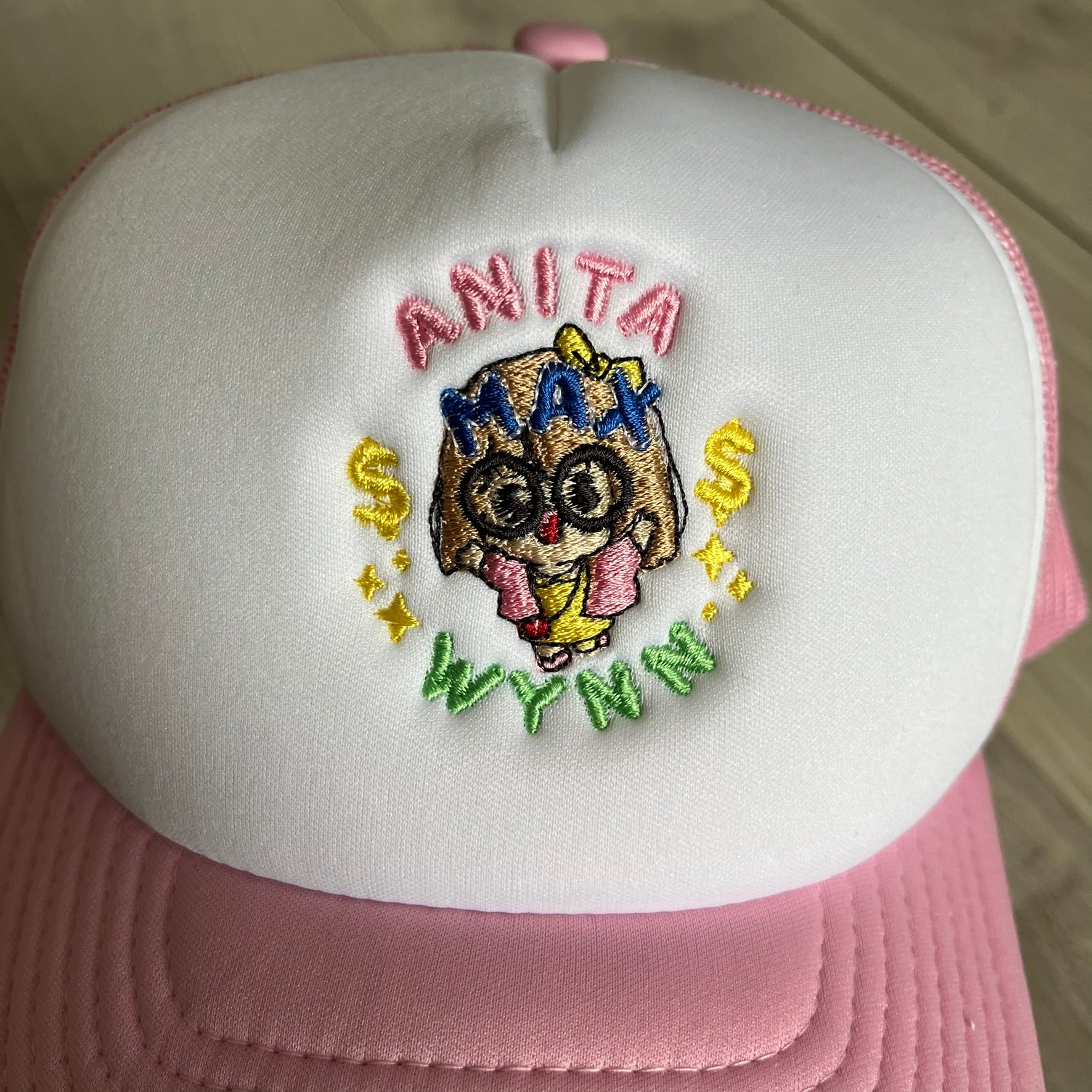 The Drake Anita Max Wynn Hat Punk is a trucker hat that pays tribute to hip-hop artist Drake’s alter ego, Anita Max Wynn. Pink sides and cap, white front.
The hat was inspired by Drake’s love of gambling and his need to get a "max win." The details include a collection of graphics on the front, such as a woman with an oversized head and a pink jacket, two yellow dollar signs, "ANITA" in pink, "MAX" in Blue, and " WYNN" in green. The hat features an construction with a mesh back. A traditional snapback provi