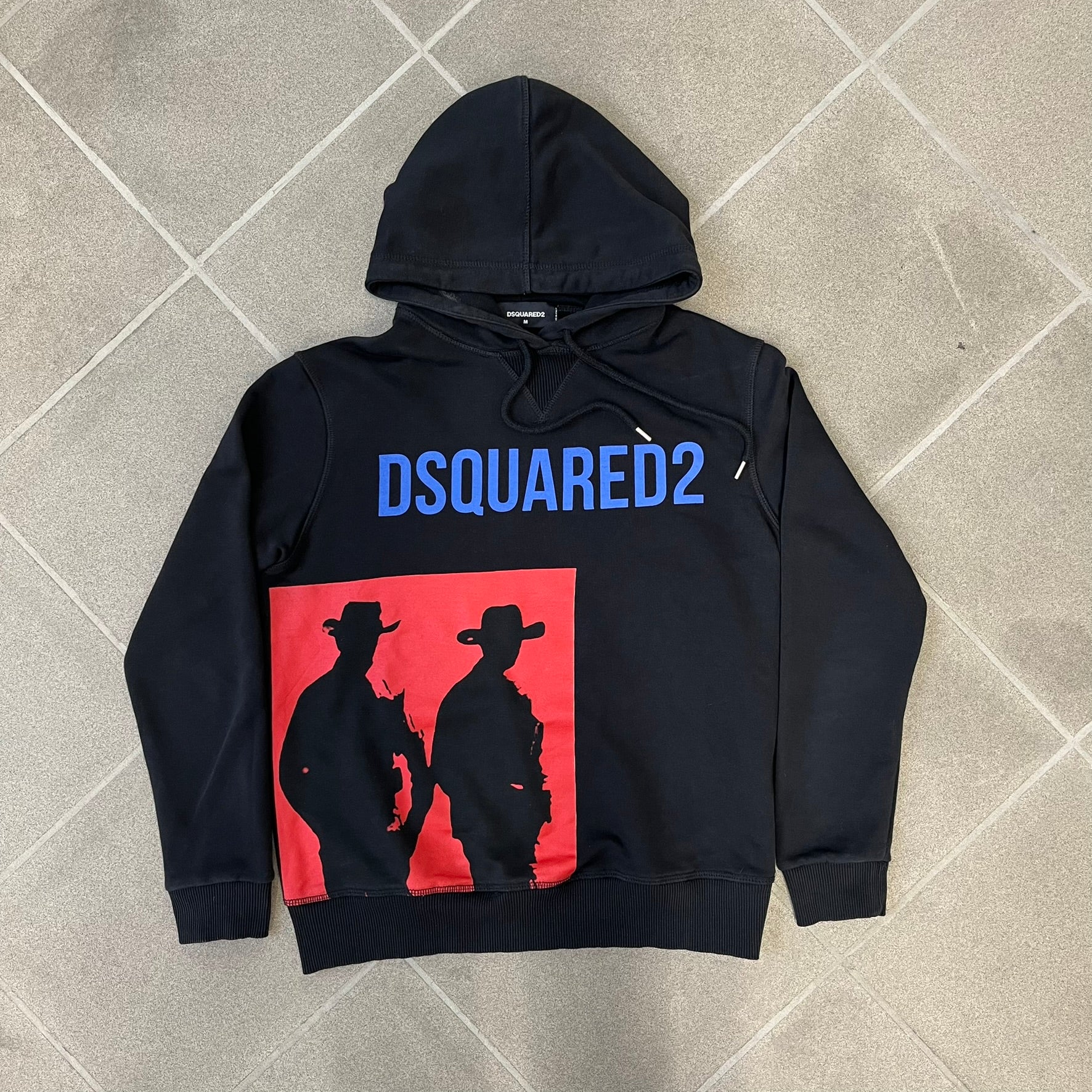 Dsquared2 2017 Western Cowboys Hoodie is a black hoodie featuring a blue Dsquared2 text logo across the upper front and a red square graphic on the lower right. Inside the square are two black cowboy figures wearing hats. The hoodie was made in Portugal.

