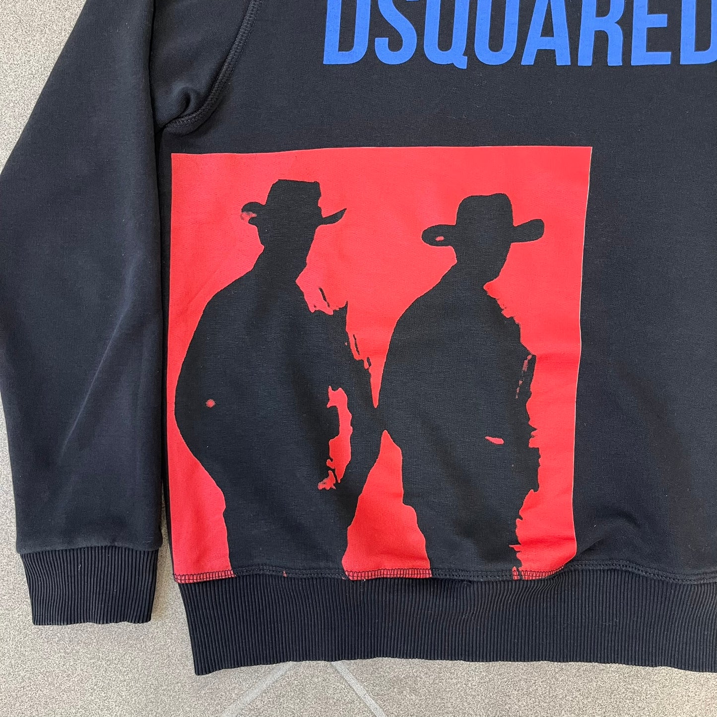 Dsquared2 2017 Western Cowboys Hoodie is a black hoodie featuring a blue Dsquared2 text logo across the upper front and a red square graphic on the lower right. Inside the square are two black cowboy figures wearing hats. The hoodie was made in Portugal.
