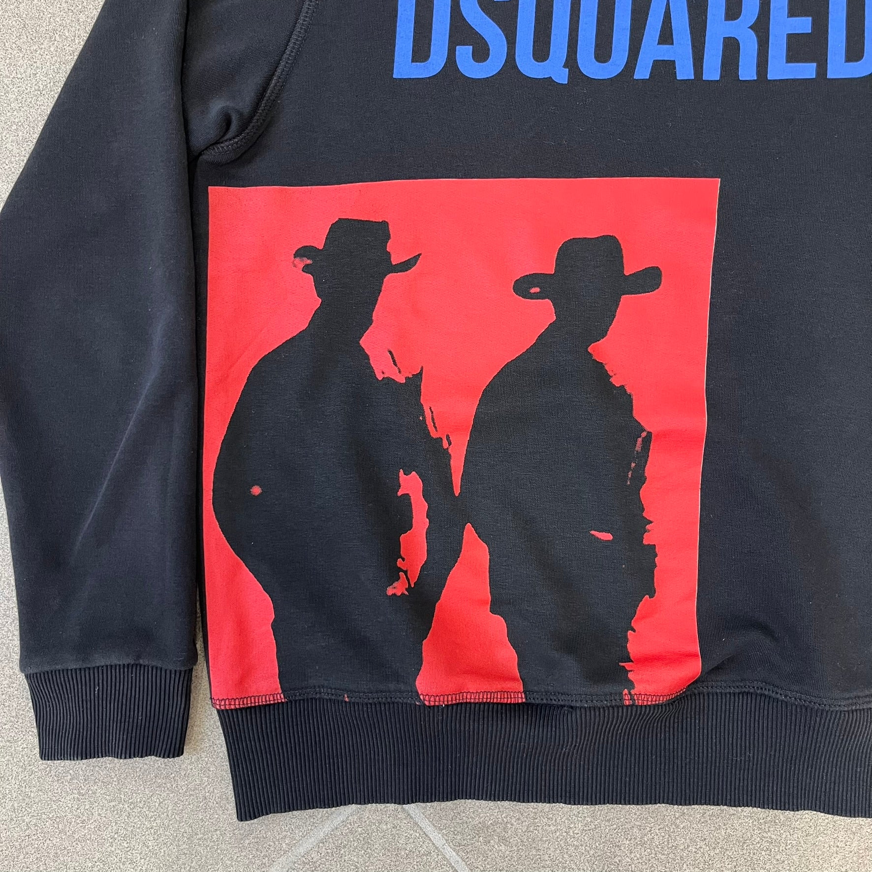 Dsquared2 2017 Western Cowboys Hoodie is a black hoodie featuring a blue Dsquared2 text logo across the upper front and a red square graphic on the lower right. Inside the square are two black cowboy figures wearing hats. The hoodie was made in Portugal.
