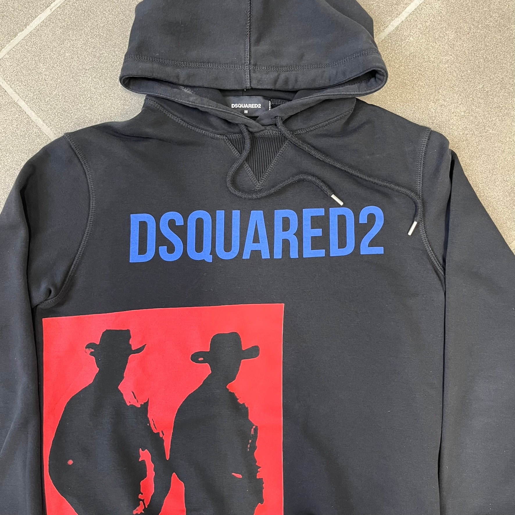 Dsquared2 2017 Western Cowboys Hoodie is a black hoodie featuring a blue Dsquared2 text logo across the upper front and a red square graphic on the lower right. Inside the square are two black cowboy figures wearing hats. The hoodie was made in Portugal.

