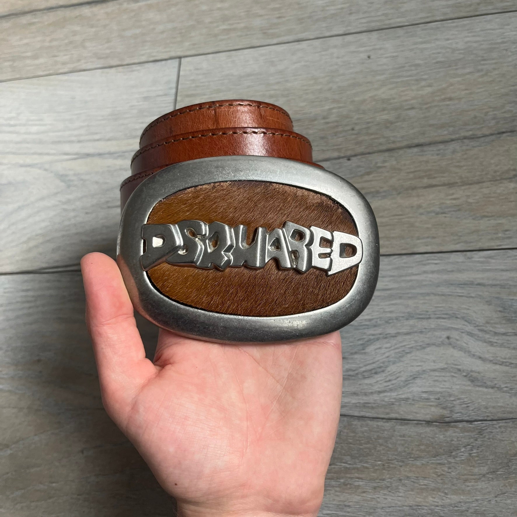 Dsquared2 Big Brown Hair Plaque Leather Belt is a leather belt featuring a large oval metal buckle with a silver-tone frame and brown short hair center. The buckle displays DSQUARED2 text in the middle, while the strap includes stamped branding and product details. Made from brown leather, the belt is size Medium.