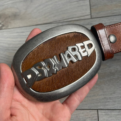 Dsquared2 Big Brown Hair Plaque Leather Belt is a leather belt featuring a large oval metal buckle with a silver-tone frame and brown short hair center. The buckle displays DSQUARED2 text in the middle, while the strap includes stamped branding and product details. Made from brown leather, the belt is size Medium.