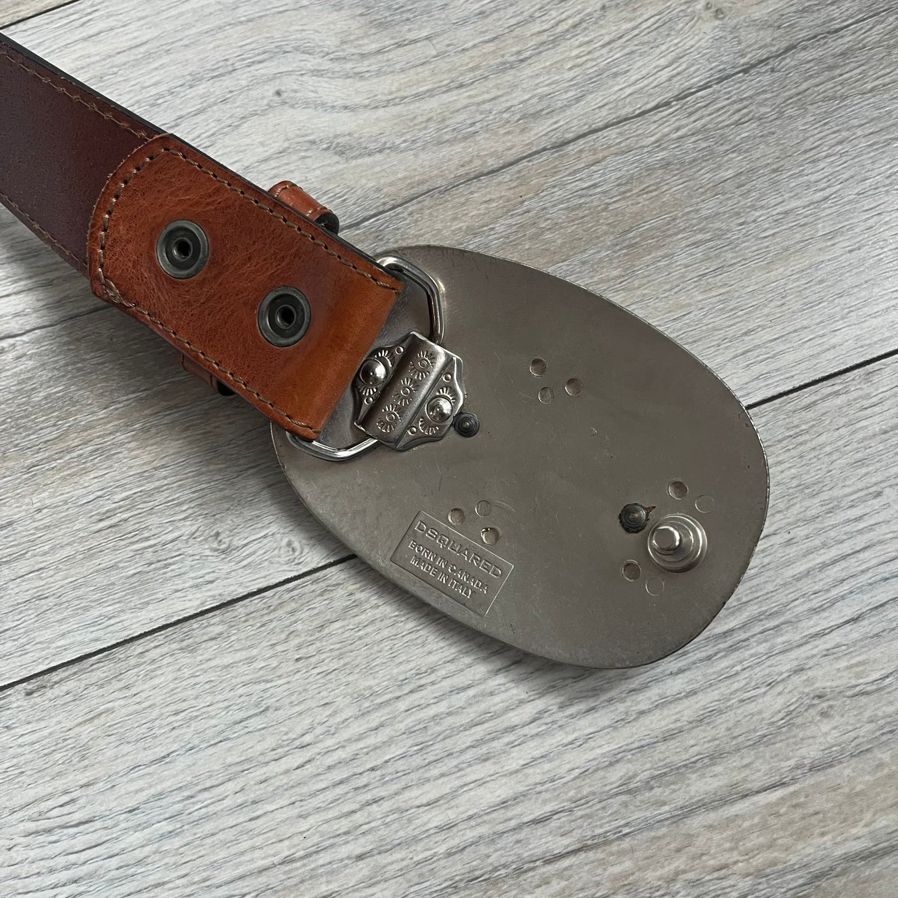 Dsquared2 Big Brown Hair Plaque Leather Belt is a leather belt featuring a large oval metal buckle with a silver-tone frame and brown short hair center. The buckle displays DSQUARED2 text in the middle, while the strap includes stamped branding and product details. Made from brown leather, the belt is size Medium.
