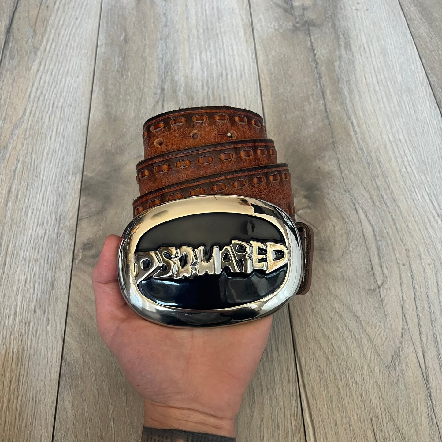 Dsquared2 Big Navy Plaque Leather Belt is a leather belt featuring a large oval metal buckle with a silver-tone frame and navy enamel center. The buckle displays DSQUARED2 text in the middle, while the strap includes stamped branding and product details. Made from brown leather, the belt is size small.