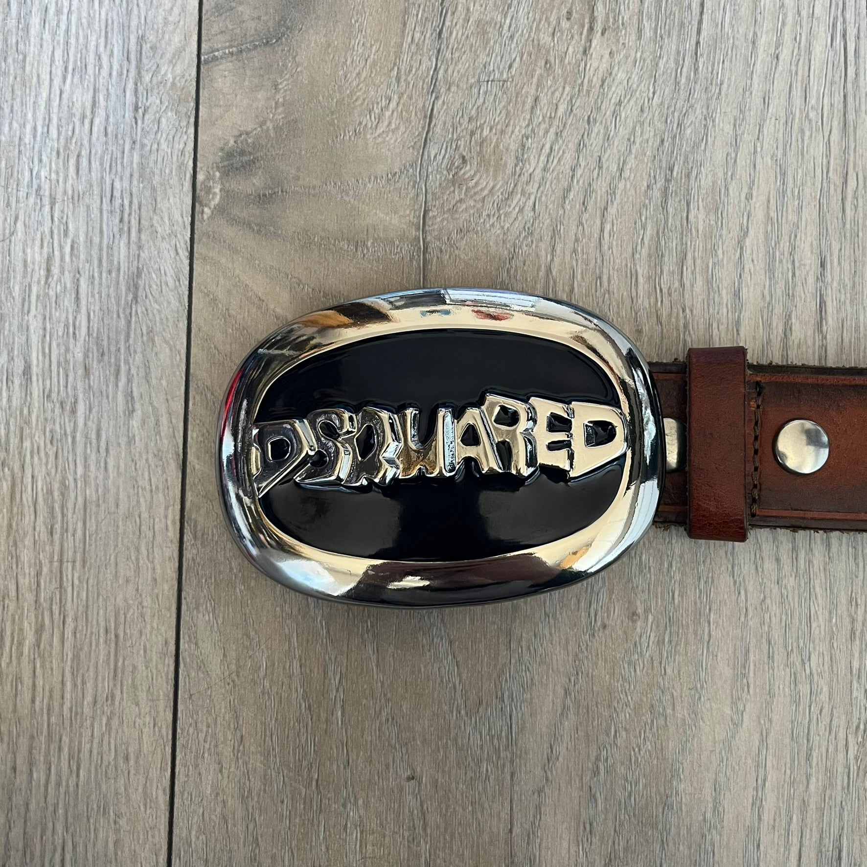 Dsquared2 Big Navy Plaque Leather Belt is a leather belt featuring a large oval metal buckle with a silver-tone frame and navy enamel center. The buckle displays DSQUARED2 text in the middle, while the strap includes stamped branding and product details. Made from brown leather, the belt is size small.