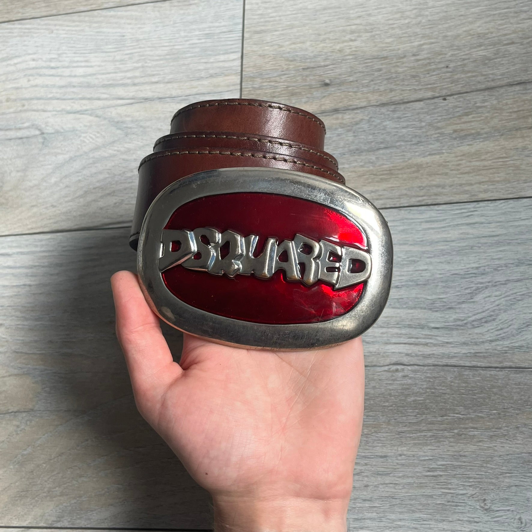 Dsquared2 Big Red Plaque Leather Belt is a leather belt featuring a large oval metal buckle with a silver-tone frame and red metallic center. The buckle displays DSQUARED2 text in the middle, while the strap includes stamped branding and product details. Made from brown leather, the belt is size small.