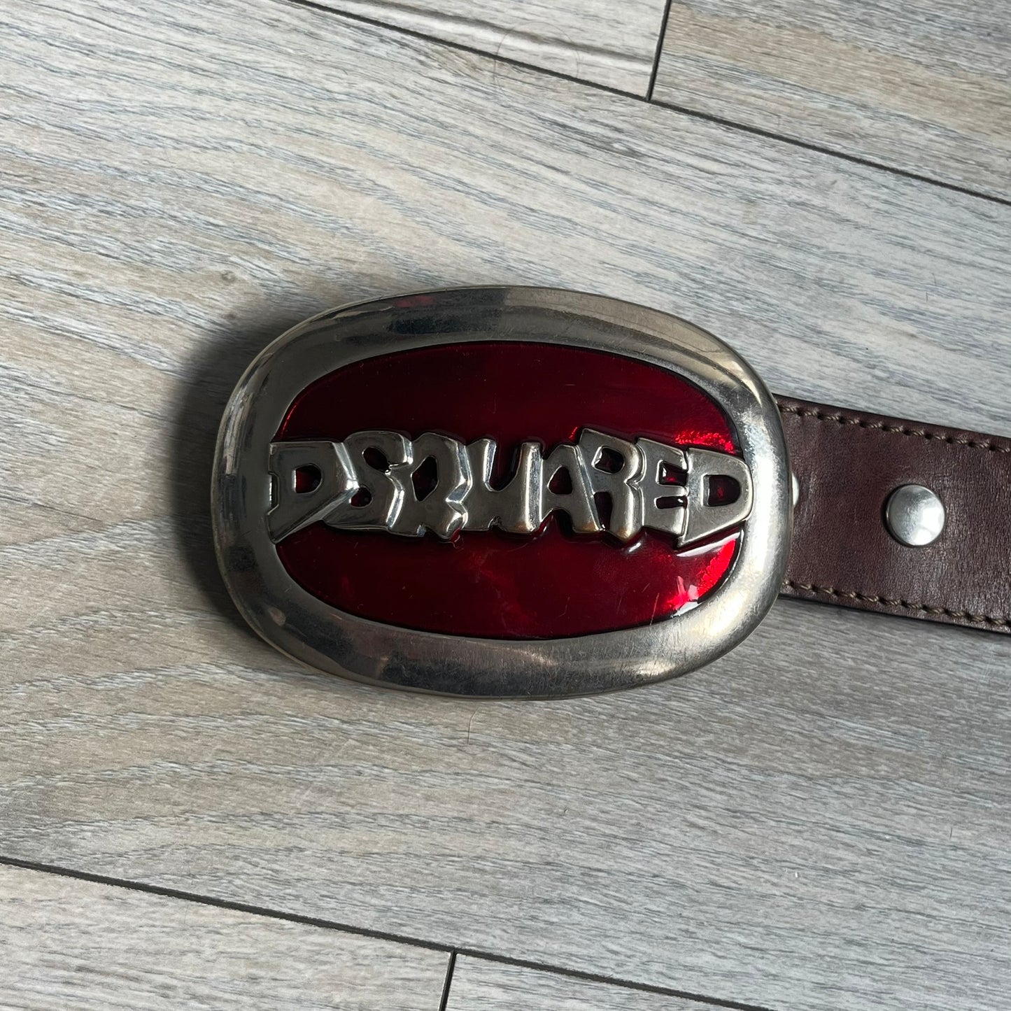 Dsquared2 Big Red Plaque Leather Belt is a leather belt featuring a large oval metal buckle with a silver-tone frame and red metallic center. The buckle displays DSQUARED2 text in the middle, while the strap includes stamped branding and product details. Made from brown leather, the belt is size small.