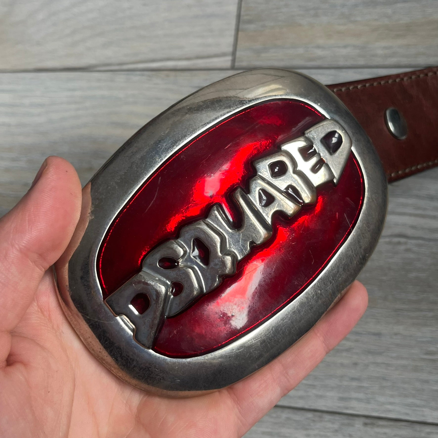 Dsquared2 Big Red Plaque Leather Belt is a leather belt featuring a large oval metal buckle with a silver-tone frame and red metallic center. The buckle displays DSQUARED2 text in the middle, while the strap includes stamped branding and product details. Made from brown leather, the belt is size small.