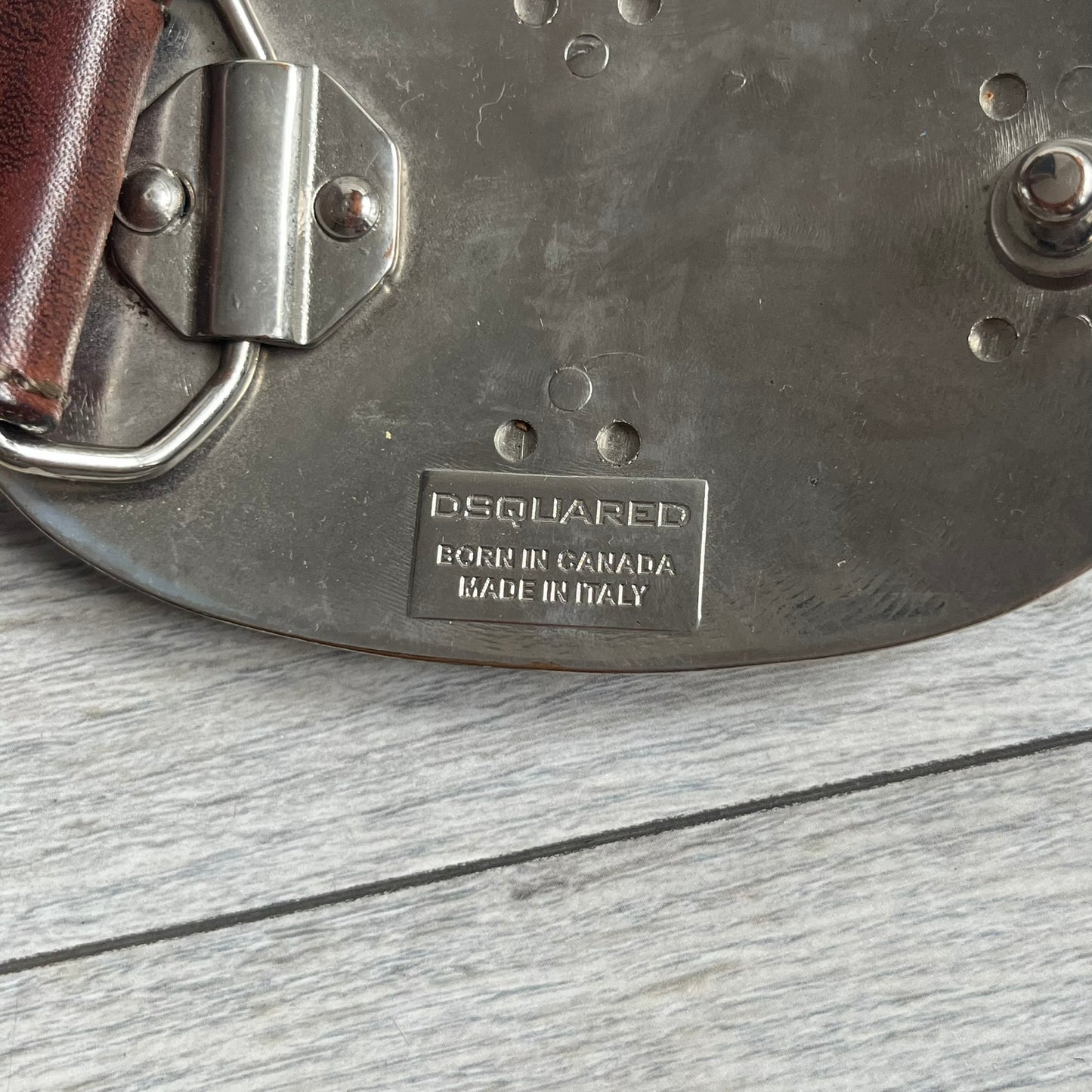 Dsquared2 Big Red Plaque Leather Belt is a leather belt featuring a large oval metal buckle with a silver-tone frame and red metallic center. The buckle displays DSQUARED2 text in the middle, while the strap includes stamped branding and product details. Made from brown leather, the belt is size small.
