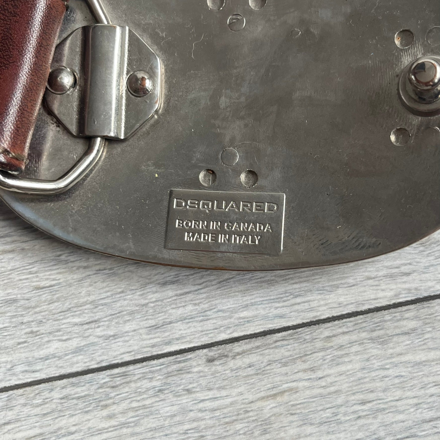 Dsquared2 Big Red Plaque Leather Belt is a leather belt featuring a large oval metal buckle with a silver-tone frame and red metallic center. The buckle displays DSQUARED2 text in the middle, while the strap includes stamped branding and product details. Made from brown leather, the belt is size small.