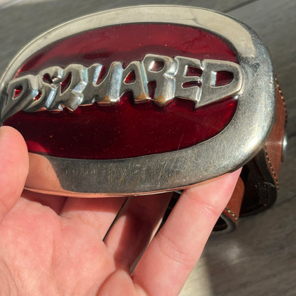 Dsquared2 Big Red Plaque Leather Belt is a leather belt featuring a large oval metal buckle with a silver-tone frame and red metallic center. The buckle displays DSQUARED2 text in the middle, while the strap includes stamped branding and product details. Made from brown leather, the belt is size small.