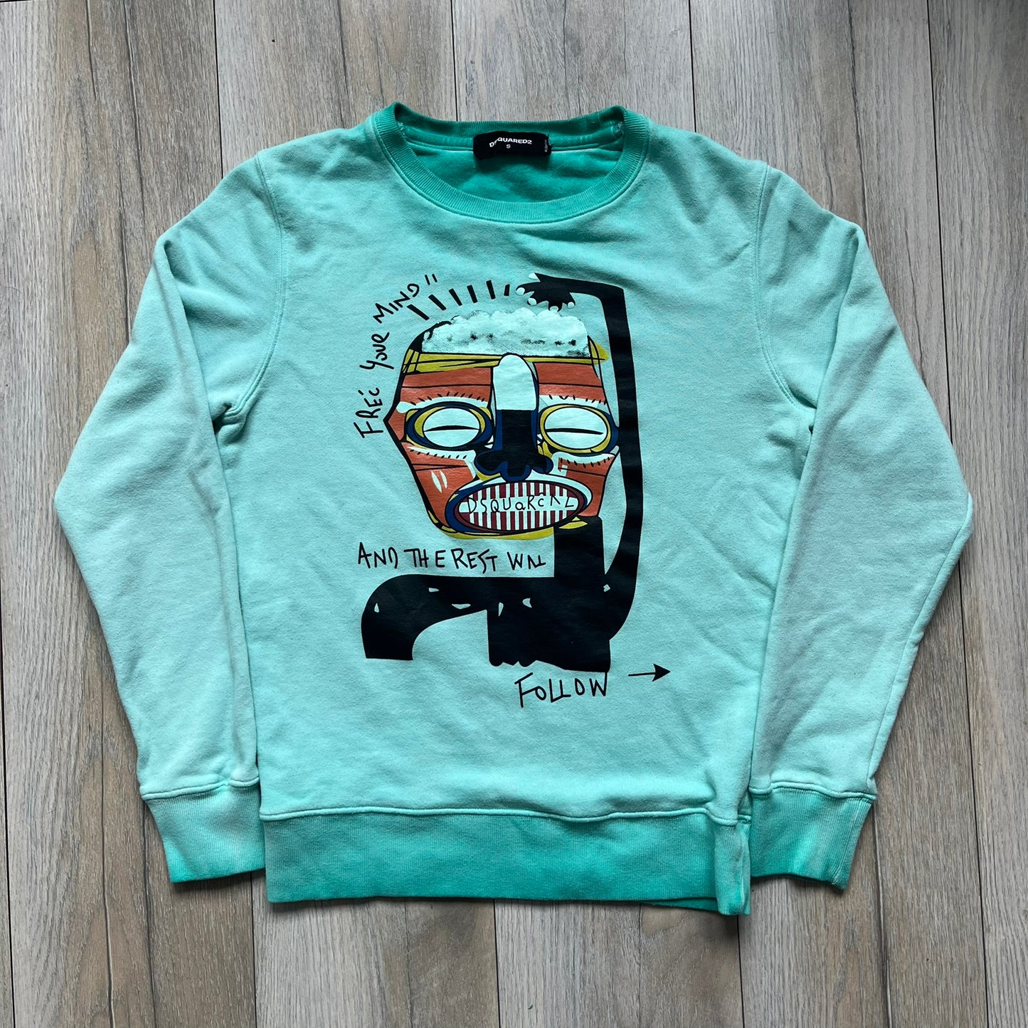 Dsquared2 SS2015 “Free Your Mind” Crewneck is a turquoise cotton sweatshirt with a washed, faded finish and tonal variation across the fabric. The front features a black Basquiat-inspired figure with “Free Your Mind and the Rest Will Follow” text in a contrasting design. The piece is made in Italy and includes darker tonal hems with a lighter center fade for a worn effect.&nbsp;