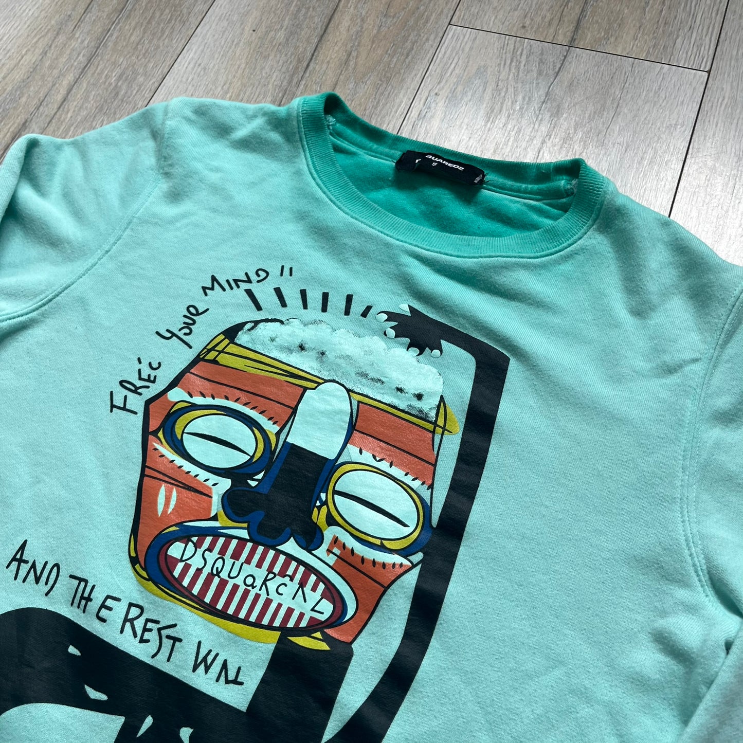 Dsquared2 SS2015 “Free Your Mind” Crewneck is a turquoise cotton sweatshirt with a washed, faded finish and tonal variation across the fabric. The front features a black Basquiat-inspired figure with “Free Your Mind and the Rest Will Follow” text in a contrasting design. The piece is made in Italy and includes darker tonal hems with a lighter center fade for a worn effect.&nbsp;