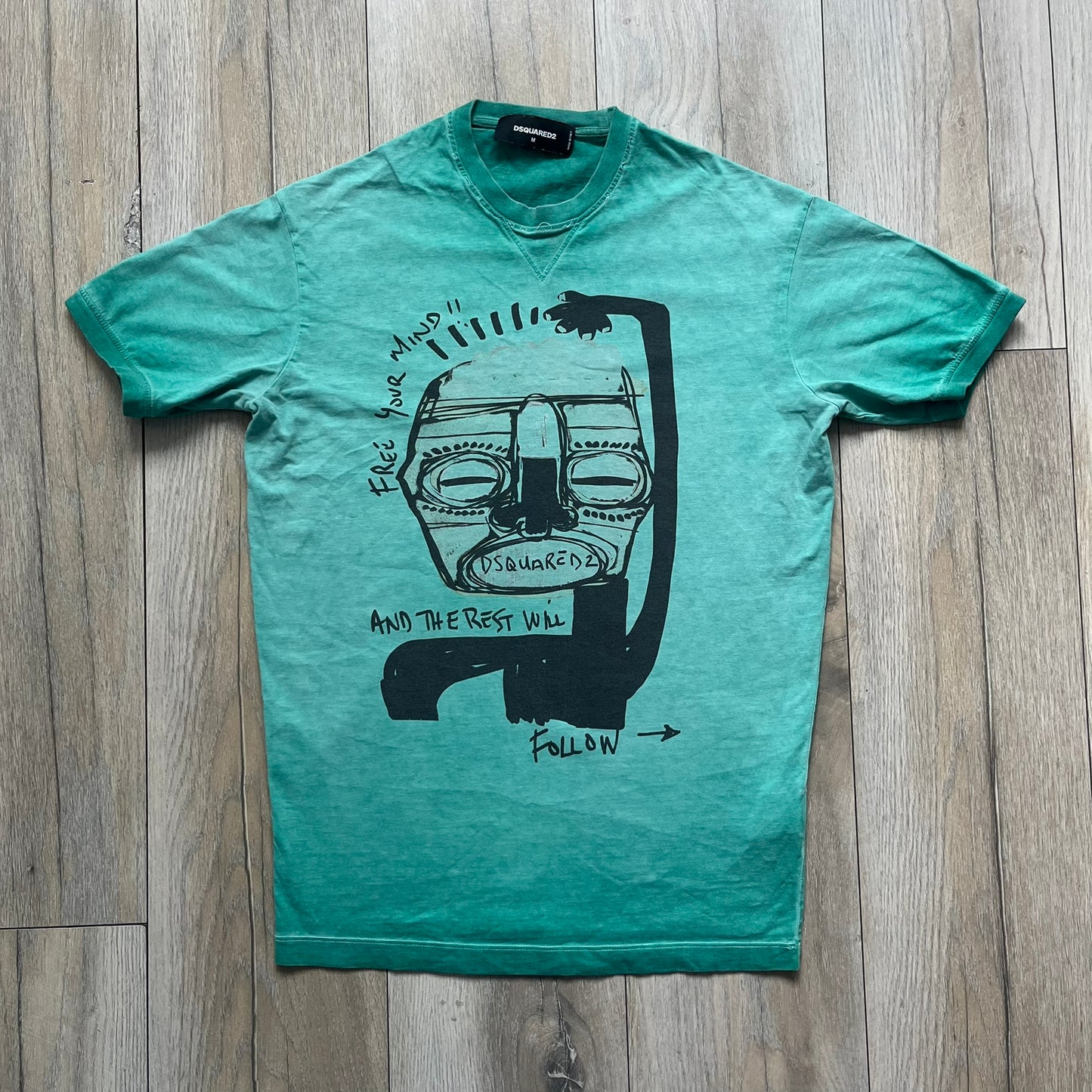 Dsquared2 SS2015 “Free Your Mind” T-shirt is a turquoise cotton tee with a washed, faded finish and tonal variation across the fabric. The front features a black Basquiat-inspired figure with “Free Your Mind and the Rest Will Follow” text in a contrasting design. The piece is made in Italy and includes darker tonal hems with a lighter center fade for a worn effect.